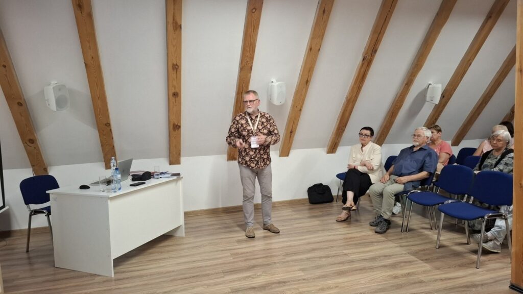 Prelegent przemawiający do zgromadzonej publiczności, uczestnicy siedzący w rzędach krzeseł w sali konferencyjnej (kliknięcie powiększy zdjęcie