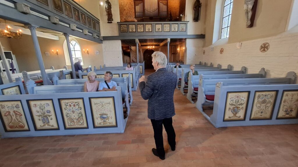 Prowadzący opowiada o historii kościoła, w ławkach kościelnych siedzą ludzie (kliknięcie powiększy zdjęcie)