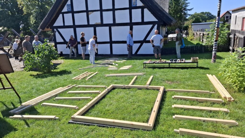 Elementy drewnianej konstrukcji rozłożone na trawie, w tle tradycyjny budynek szachulcowy, uczestnicy przygotowujący się do składania konstrukcji (kliknięcie powiększy zdjęcie)