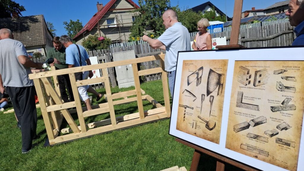 Plansza z historycznymi rysunkami technicznymi drewnianych połączeń, w tle uczestnicy montujący konstrukcję (kliknięcie powiększy zdjęcie)