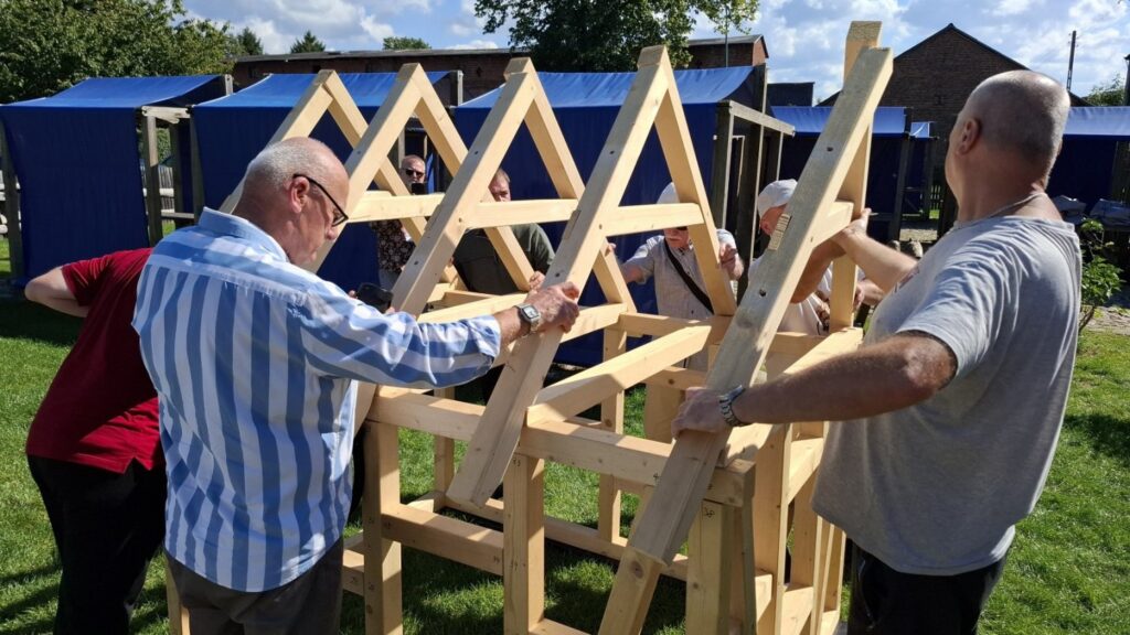 Uczestnicy podnoszą i dopasowują elementy dachu drewnianej konstrukcji (kliknięcie powiększy zdjęcie)