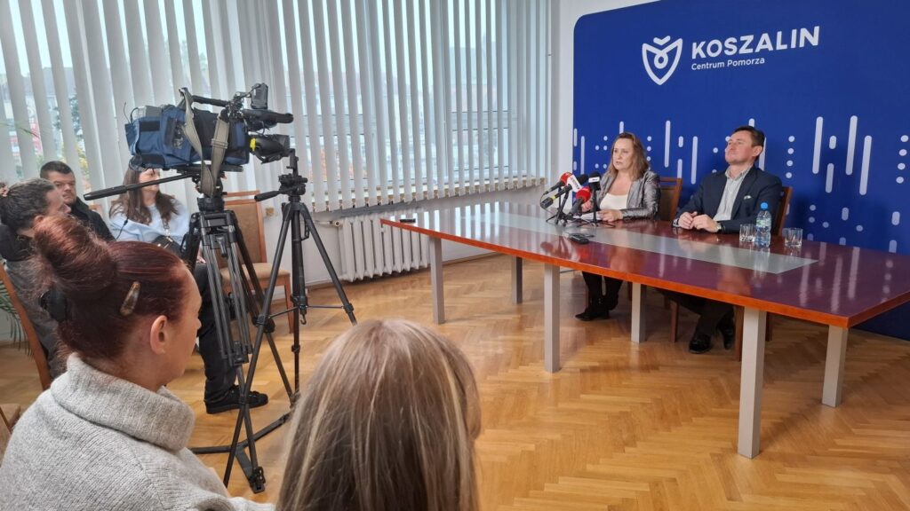 Dwie osoby siedzą przy stole, na którym stoją mikrofony. Po lewej - pracownicy mediów i kamera na statywie (kliknięcie spowoduje powiększenie zdjęcia).