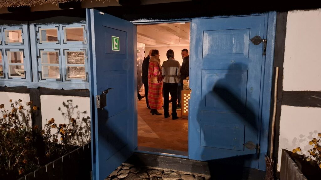 Otwarte niebieskie drzwi tradycyjnego budynku, przez które widać wnętrze z uczestnikami wernisażu wystawy (kliknięcie powiększy zdjęcie)