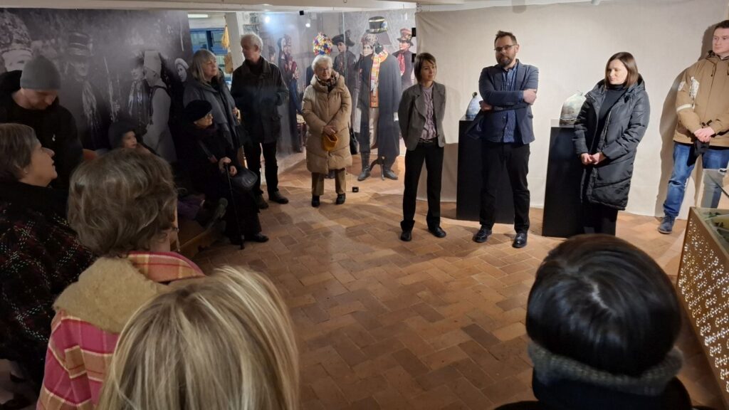 Grupa osób stojących w kręgu we wnętrzu muzeum, słuchających oprowadzania po wystawie etnograficznej (kliknięcie powiększy zdjęcie)
