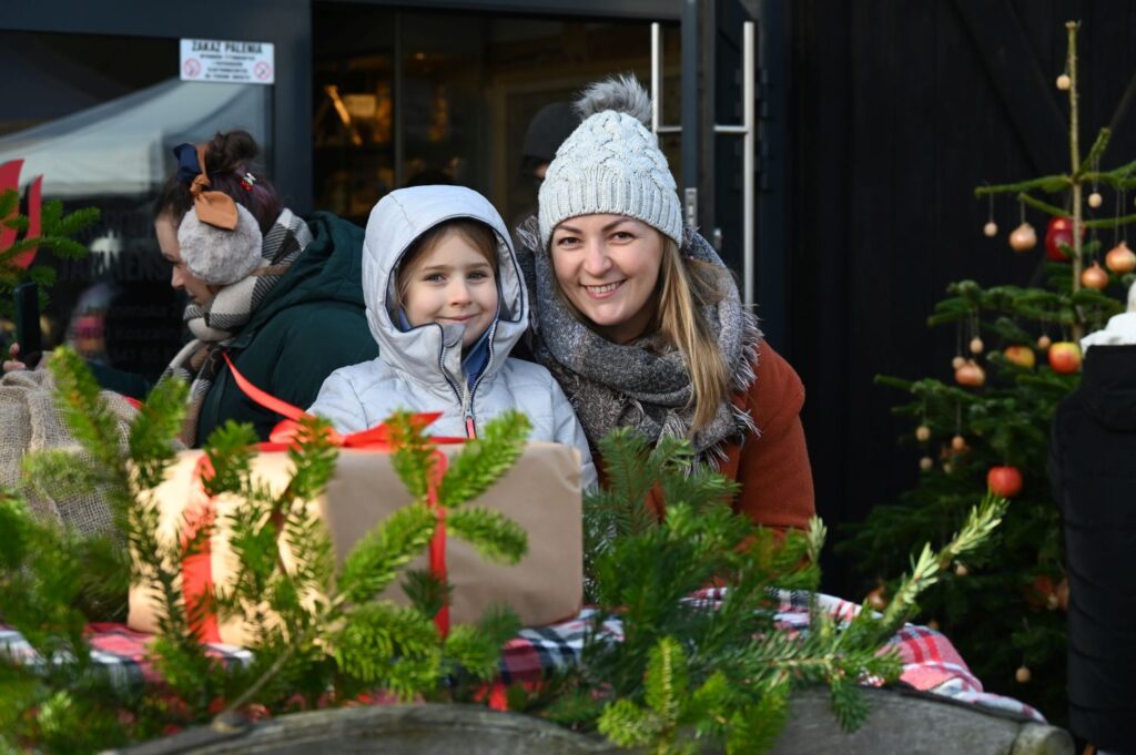 Kobieta i dziecko uśmiechają się przy stoisku jarmarcznym, na którym leżą świąteczne dekoracje i zapakowany prezent (kliknięcie powiększy zdjęcie).