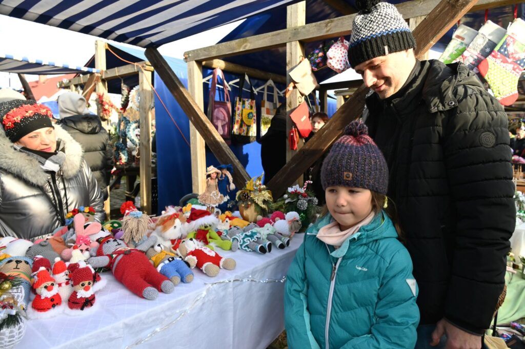 Rodzina ogląda kolorowe lalki i zabawki rękodzielnicze na stoisku jarmarcznym, dziecko stoi przy stole z ekspozycją (kliknięcie powiększy zdjęcie).