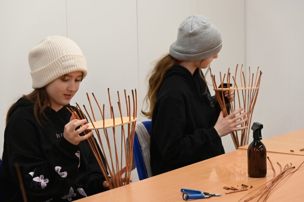 Dwie dziewczynki siedzą przy stole i wyplatają dekoracje z cienkich gałązek podczas warsztatów rękodzielniczych (kliknięcie powiększy zdjęcie).