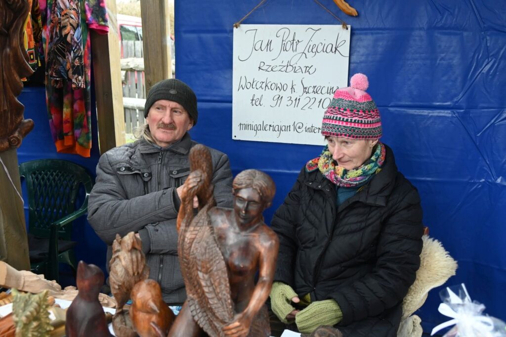 Sprzedawcy siedzą przy stoisku z drewnianymi rzeźbami i figurami wystawionymi na sprzedaż (kliknięcie powiększy zdjęcie).