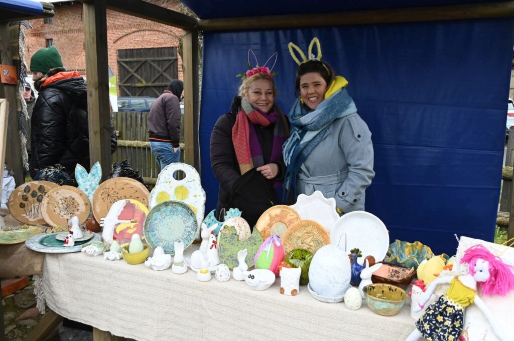 Dwie sprzedawczynie stoją za stołem z rękodziełem i ceramicznymi ozdobami wielkanocnymi (kliknięcie powiększy zdjęcie).
