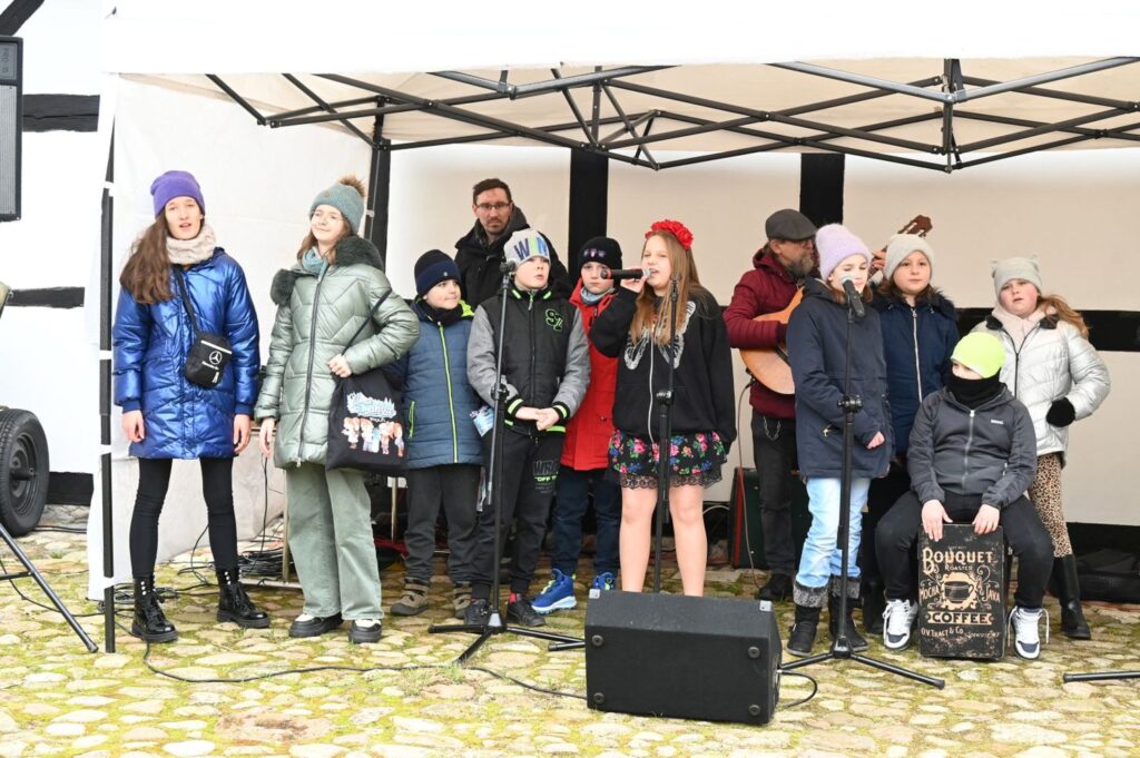 Grupa dzieci i młodzieży śpiewa do mikrofonów podczas występu pod zadaszeniem sceny (kliknięcie powiększy zdjęcie).