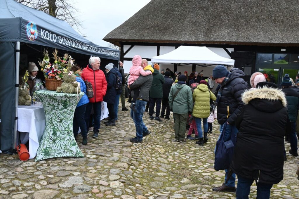 Odwiedzający stoją przy stoisku Koła Gospodyń Wiejskich ustawionym przed budynkiem krytym strzechą (kliknięcie powiększy zdjęcie).