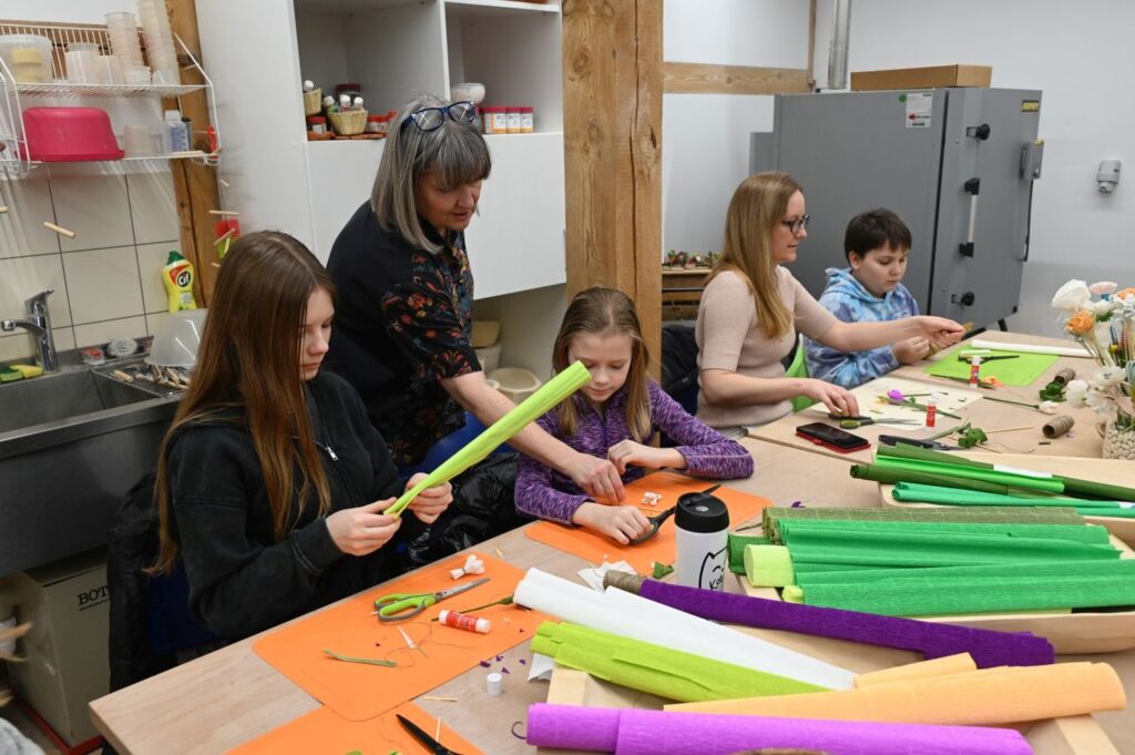 Dzieci i prowadząca siedzą przy stole podczas warsztatów plastycznych, przygotowując ozdoby z kolorowych materiałów (kliknięcie powiększy zdjęcie).