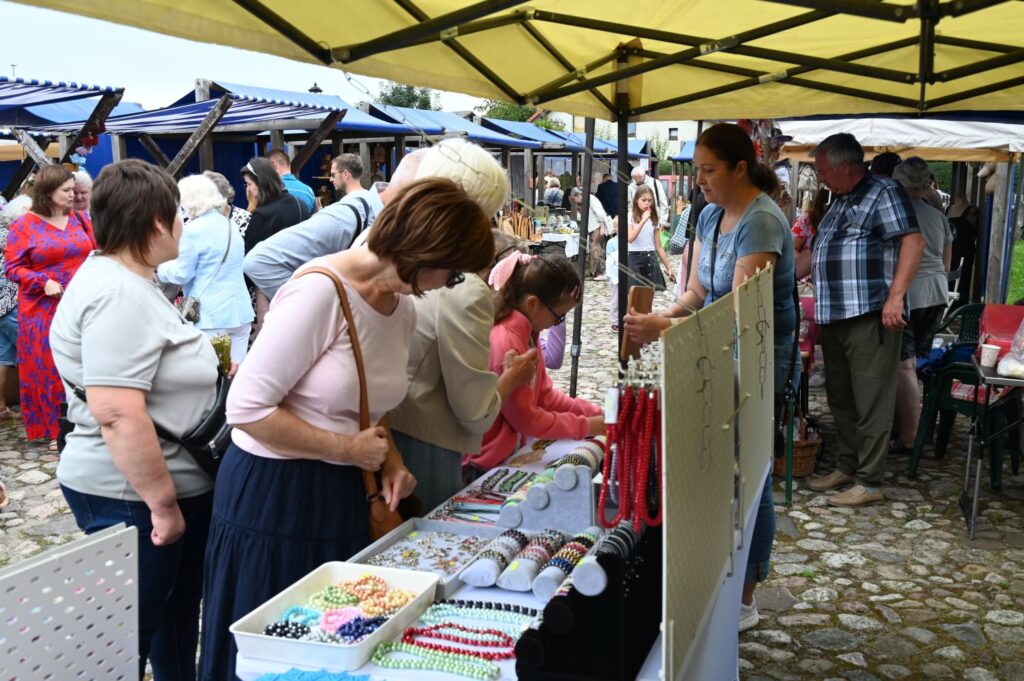 Odwiedzający oglądają biżuterię i drobne wyroby rękodzielnicze wyłożone na stoisku pod żółtym namiotem (kliknięcie powiększy zdjęcie).