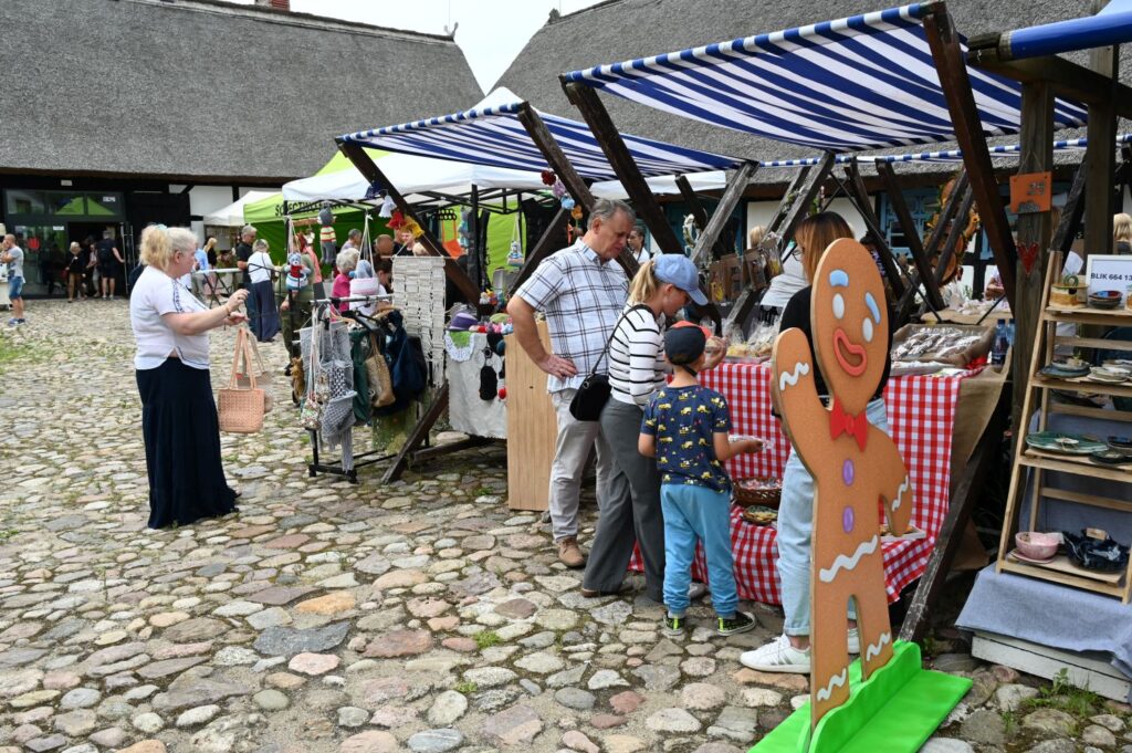 Stoisko z rękodziełem i ozdobami przy którym stoją dorośli i dziecko, a z przodu widoczna jest duża dekoracyjna postać imitująca piernika (kliknięcie powiększy zdjęcie).