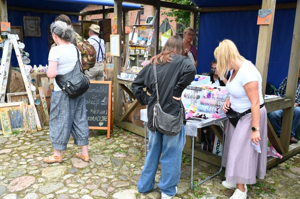 Kobiety oglądają obrazy, kartki i drobne rękodzieło na stoisku artystycznym ustawionym przy drewnianej altanie (kliknięcie powiększy zdjęcie).