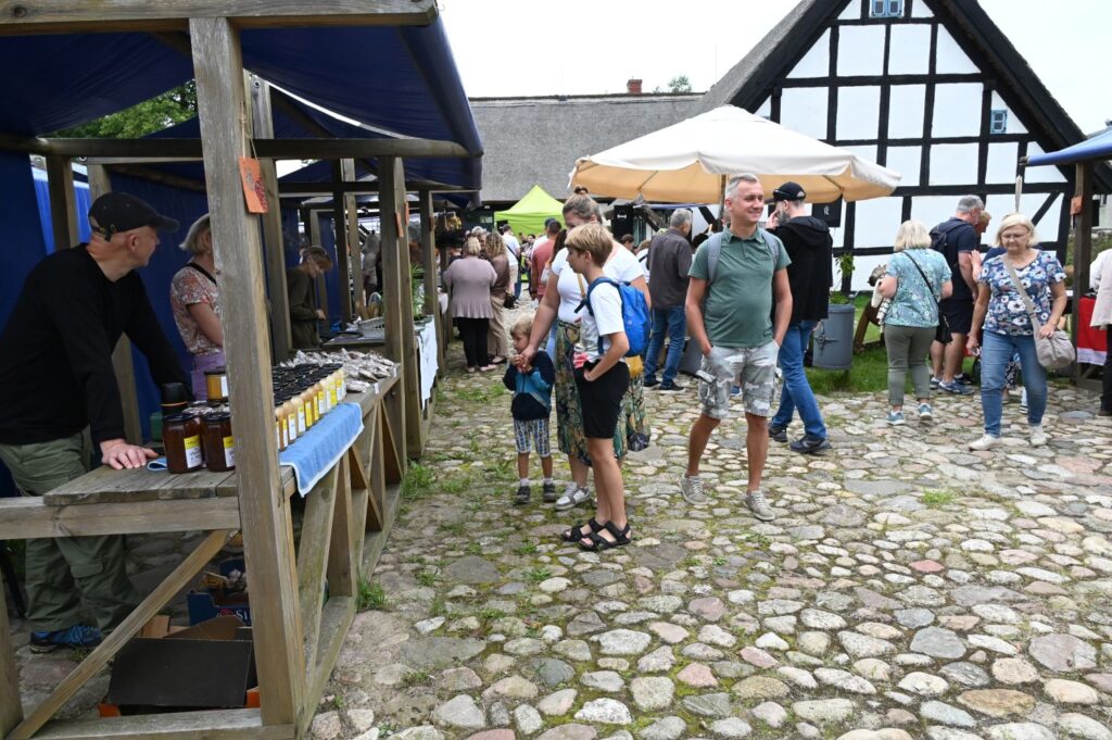 Stragany jarmarku jamneńskiego ustawione na brukowanym dziedzińcu, przy których odwiedzający oglądają słoiki z przetworami i rozmawiają ze sprzedawcą (kliknięcie powiększy zdjęcie).