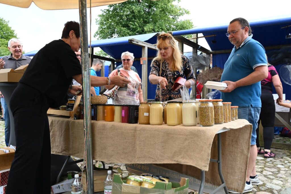 Stoisko z miodami i produktami pszczelimi, przy którym sprzedawczyni rozmawia z klientami oglądającymi słoiki z miodem (kliknięcie powiększy zdjęcie).