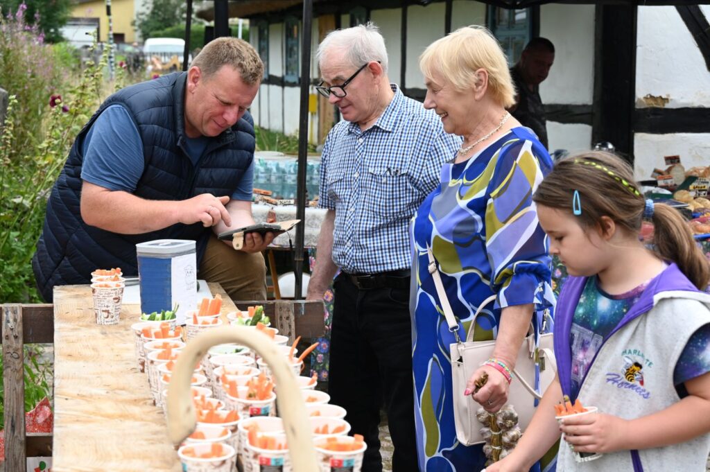 Odwiedzający stoją przy stole z potrawą dla owiec i rozmawiają z mężczyzną, który pokazuje coś na swoim telefonie (kliknięcie powiększy zdjęcie).