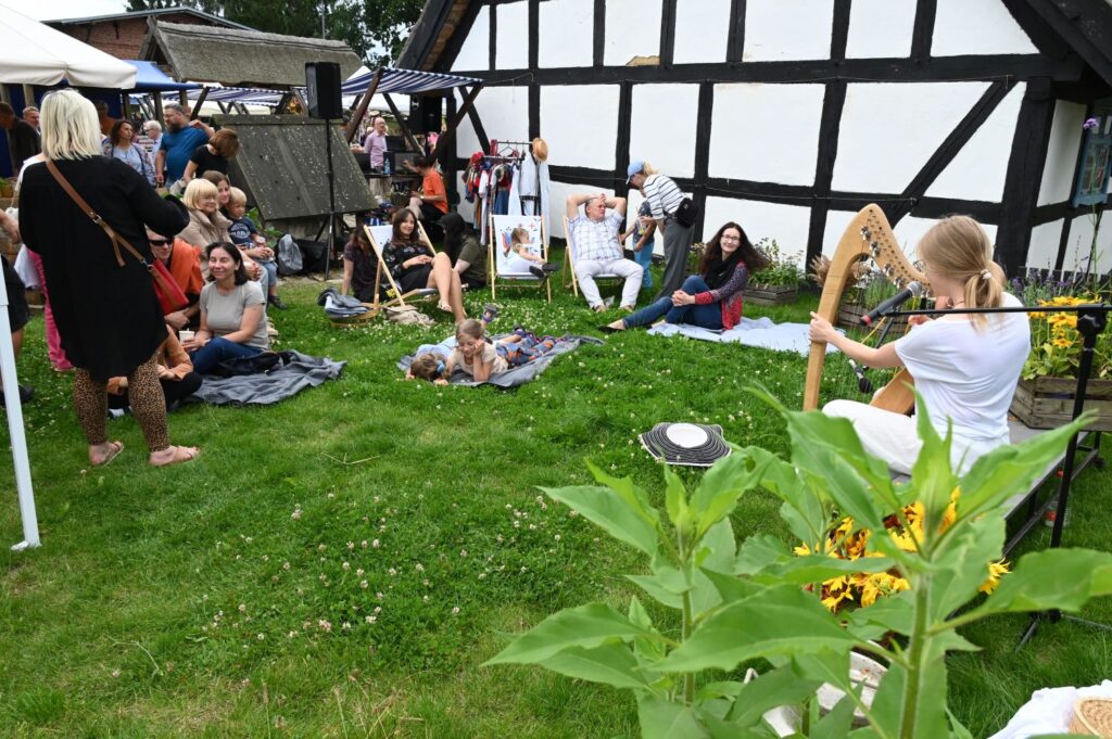 Publiczność siedzi na trawie i słucha koncertu harfowego odbywającego się na niewielkiej scenie na terenie skansenu (kliknięcie powiększy zdjęcie).