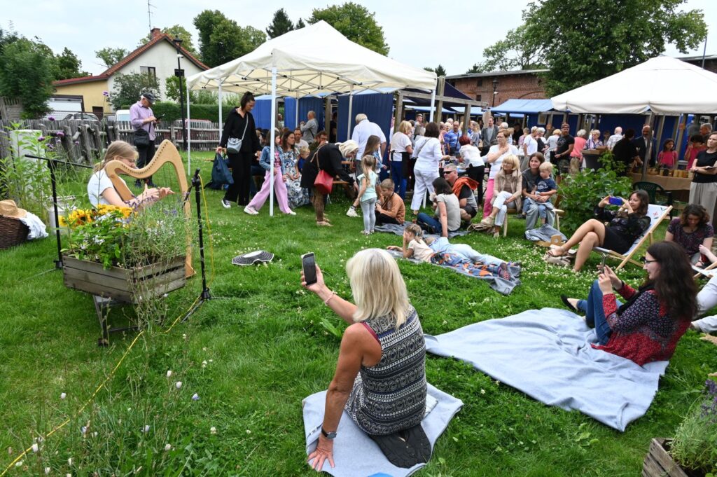 Publiczność siedzi na kocach i na trawie podczas koncertu harfowego odbywającego się na dziedzińcu jarmarku jamneńskiego (kliknięcie powiększy zdjęcie).