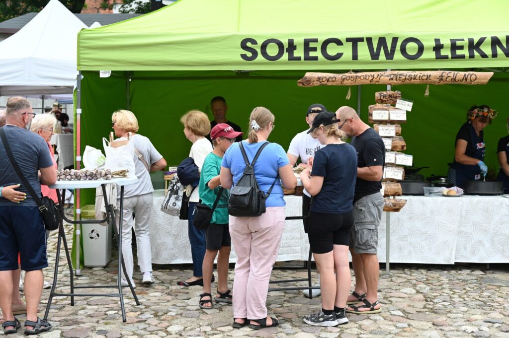 Odwiedzający stoją przy stoisku gastronomicznym pod zielonym namiotem z napisem „Sołectwo Łekno” (kliknięcie powiększy zdjęcie).