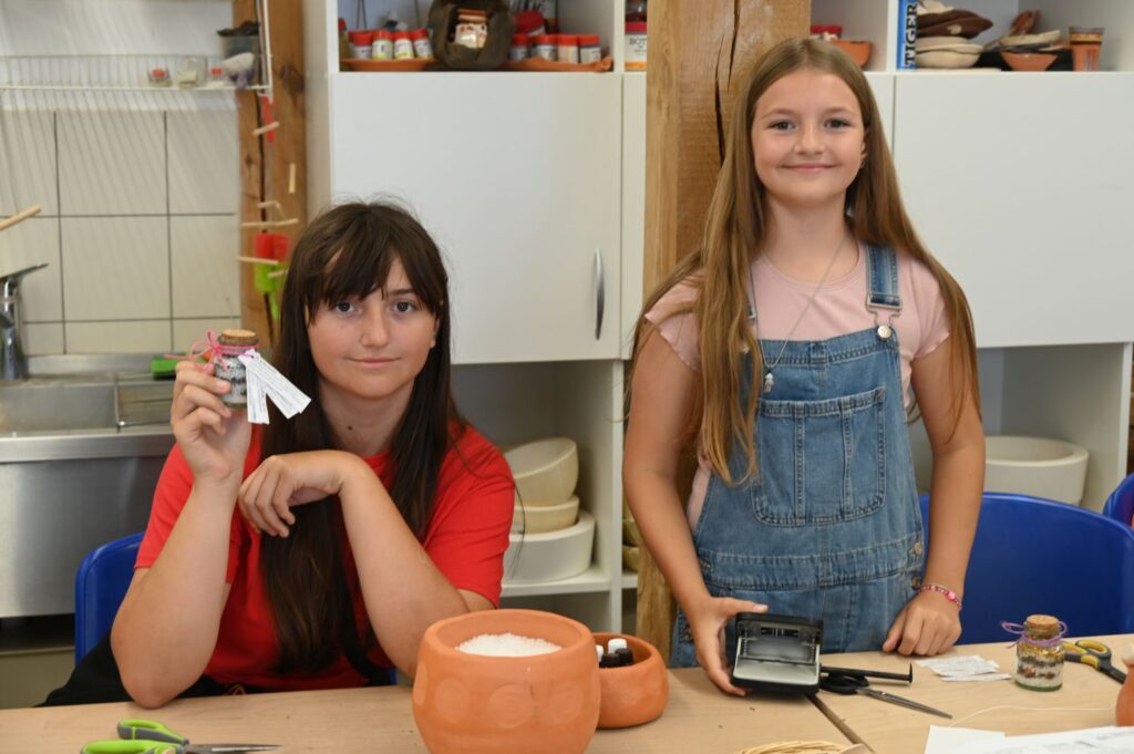 Dwie dziewczynki przy stole warsztatowym i prezentują przygotowane przez siebie słoiczki z mieszankami ziół (kliknięcie powiększy zdjęcie).