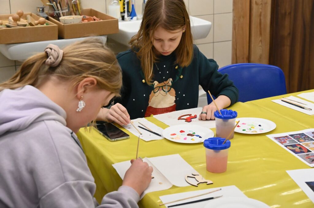 Dwie dziewczynki skupione na malowaniu świątecznych ilustracji przy stole warsztatowym (kliknięcie powiększy zdjęcie).