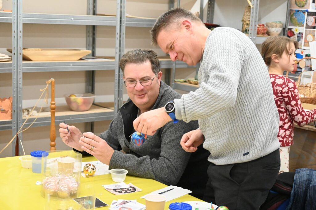 Dwóch mężczyzn uczestniczy w warsztatach plastycznych przy stole z materiałami plastycznymi, za nimi dziewczynka, która również wykonuje świąteczne ozdoby (kliknięcie powiększy zdjęcie).