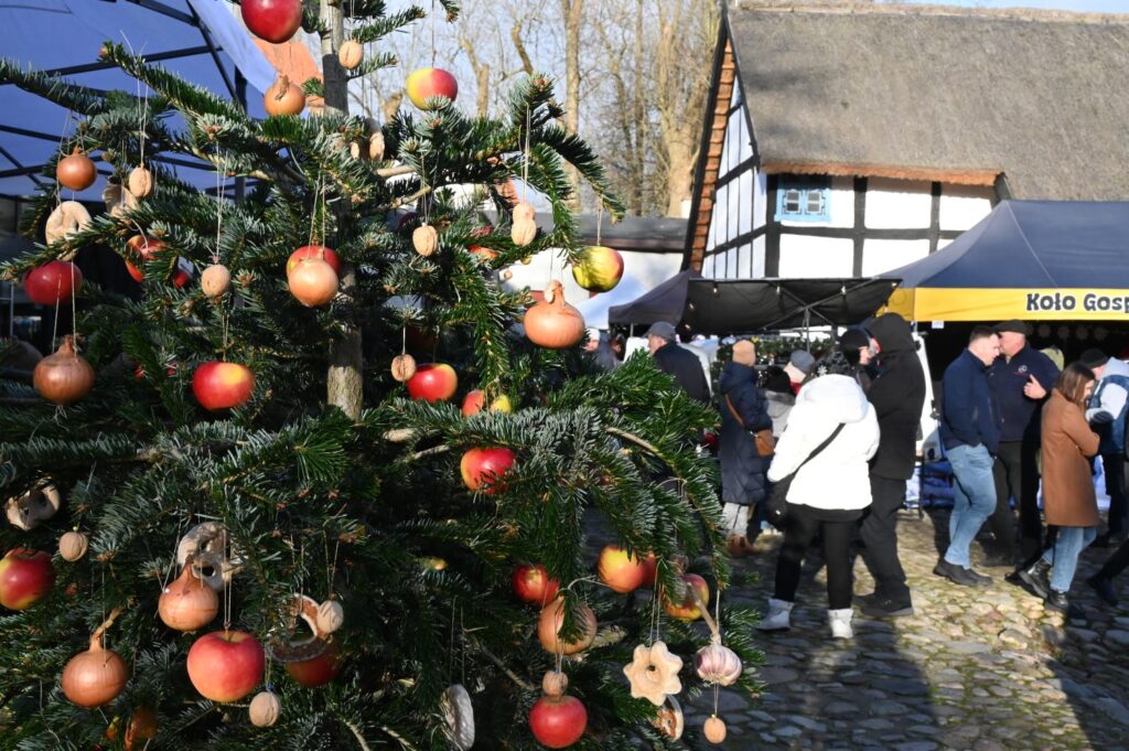Ozdobiona choinka z czerwonymi i złotymi bombkami na tle stoisk jarmarku oraz spacerujących ludzi (kliknięcie powiększy zdjęcie).