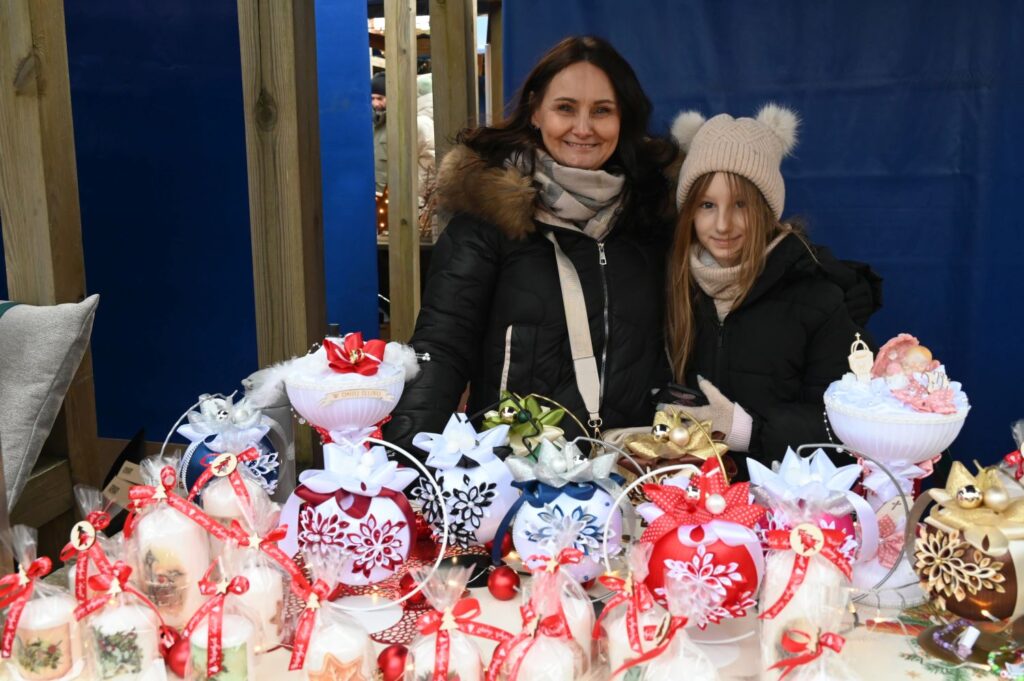 Kobieta i dziewczynka stoją za stołem z ręcznie wykonanymi ozdobami świątecznymi i uśmiechają się do zdjęcia (kliknięcie powiększy zdjęcie).