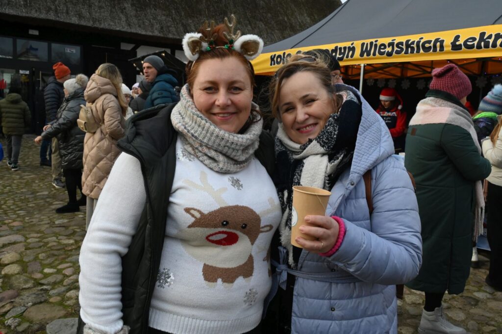 Dwie kobiety uśmiechają się do zdjęcia, trzymając kubki z napojami na tle jarmarku (kliknięcie powiększy zdjęcie).