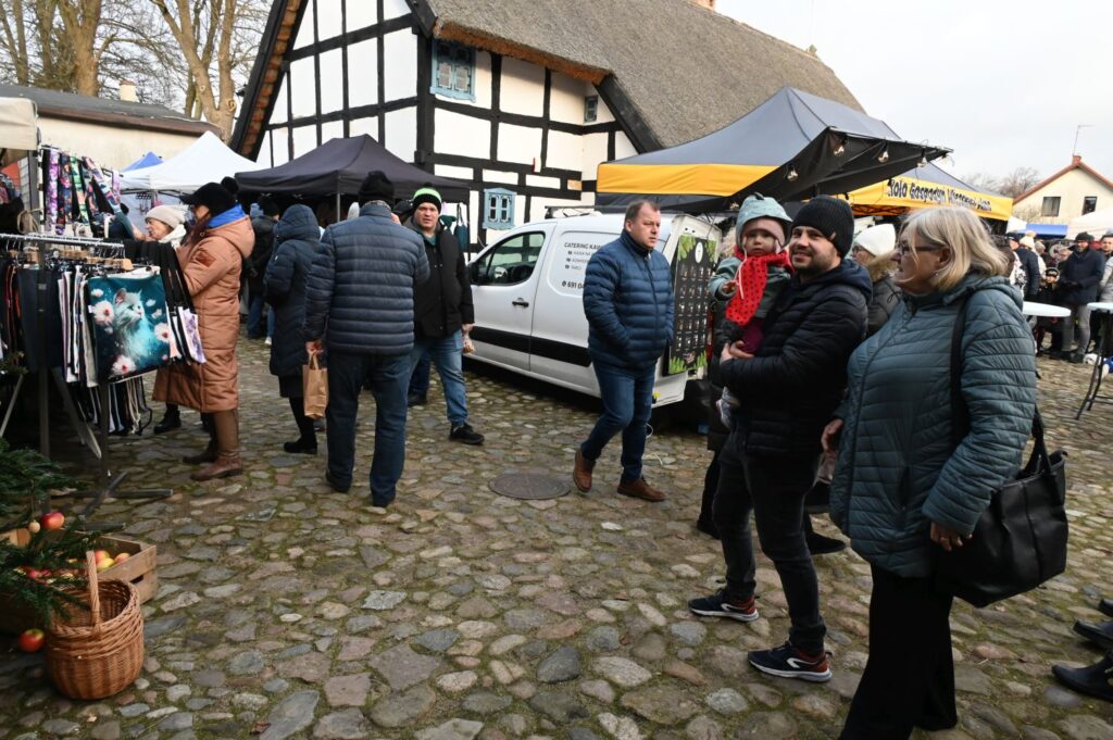 Odwiedzający spacerują między stoiskami jarmarku na brukowanym placu wśród zabudowy skansenu (kliknięcie powiększy zdjęcie).