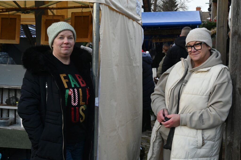 Dwie kobiety stoją przy stoisku jarmarku i patrzą w stronę aparatu (kliknięcie powiększy zdjęcie).