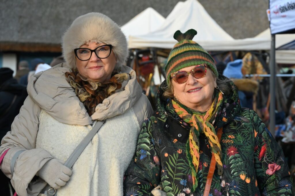 Dwie starsze kobiety w zimowych ubraniach uśmiechają się do zdjęcia na tle stoisk jarmarku (kliknięcie powiększy zdjęcie).