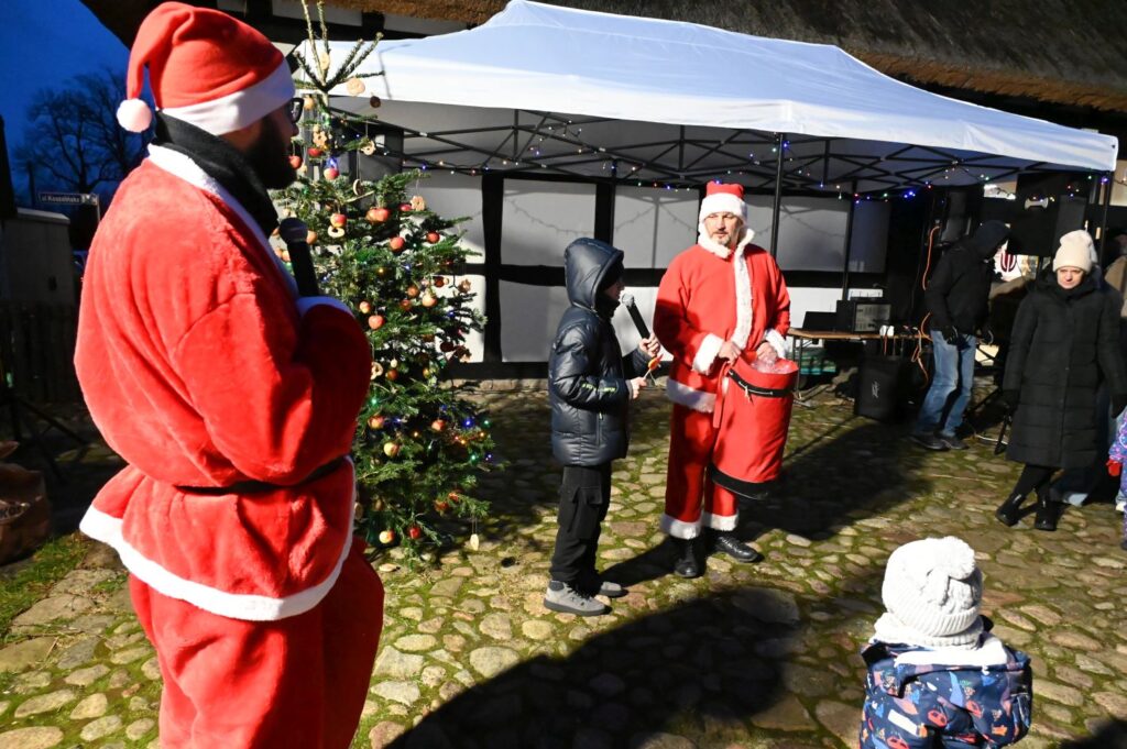 Mikołaj stoi przy udekorowanej choince i rozmawia z dziećmi podczas wydarzenia (kliknięcie powiększy zdjęcie).