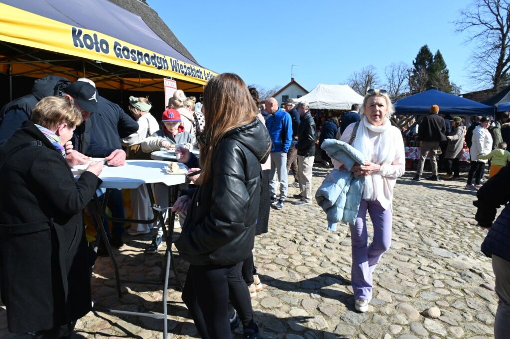 Odwiedzający stoją przy stoisku gastronomicznym podczas wielkanocnego Jarmarku Jamneńskiego na dziedzińcu zagrody (kliknięcie powiększy zdjęcie).