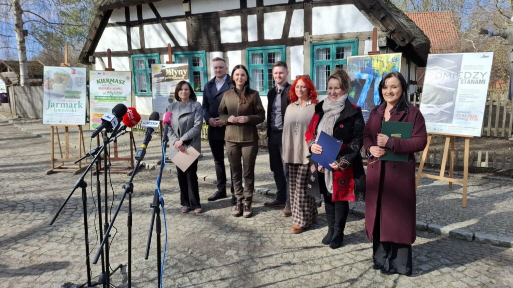 Grupa organizatorów i partnerów stoi przed budynkiem zabytkowej chałupy podczas konferencji prasowej, obok ustawione są mikrofony dla mediów oraz plansze z plakatami wydarzeń (kliknięcie powiększy zdjęcie).