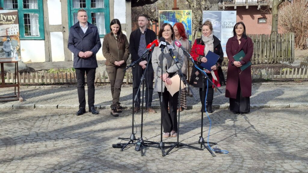 Anna Michalik przemawia do mikrofonów podczas konferencji prasowej przed budynkiem zabytkowej chałupy, a za nią stoją pozostali uczestnicy konferencji (kliknięcie powiększy zdjęcie).
