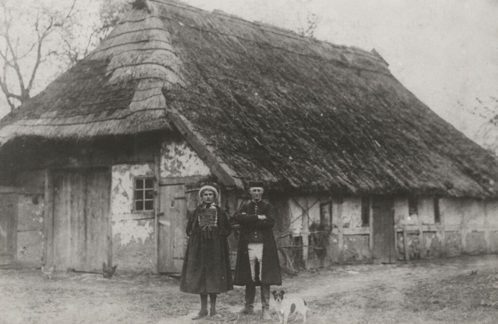 Czarno-białe archiwalne zdjęcie przedstawiające kobietę i mężczyznę stojących przed wiejską chałupą krytą strzechą, ubranych w tradycyjne stroje, u ich stóp siedzi mały pies, a w tle widoczna jest ściana budynku z małym oknem i drewnianymi drzwiami (kliknięcie powiększy zdjęcie).
