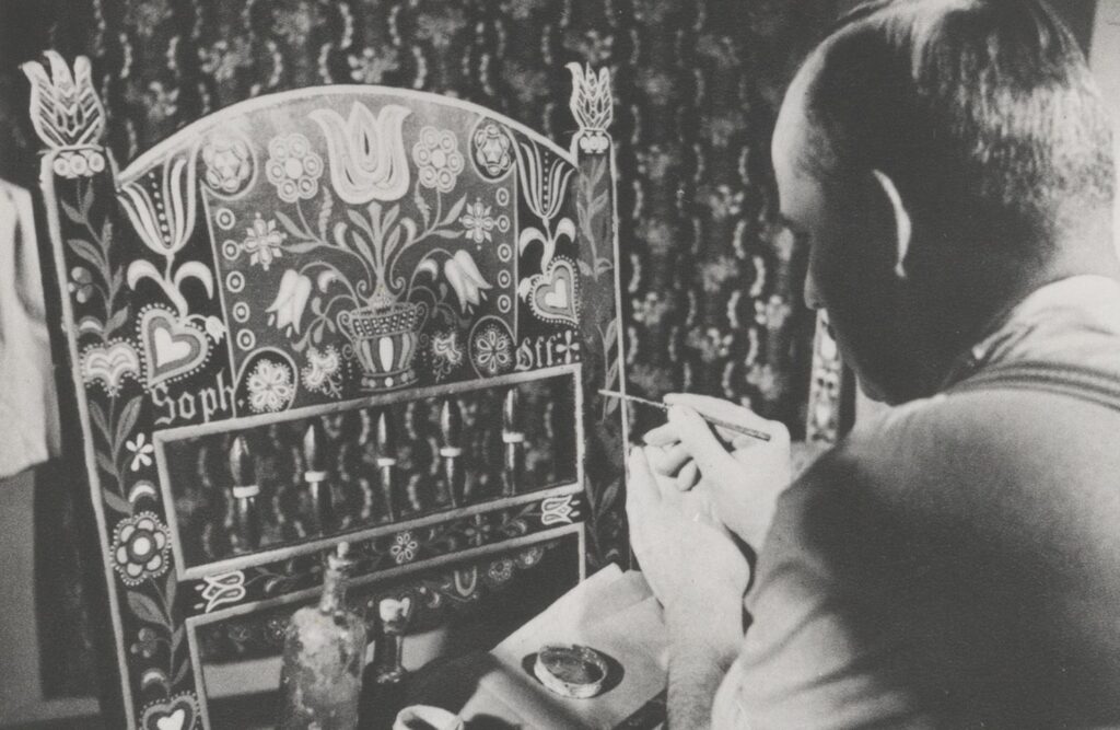 Archiwalne zdjęcie ukazujące mężczyznę malującego bogato zdobione oparcie krzesła z jamneńskimi motywami kwiatowymi i sercami, siedzącego przy stole z farbami, na tle dekoracyjnej tkaniny (kliknięcie powiększy zdjęcie).