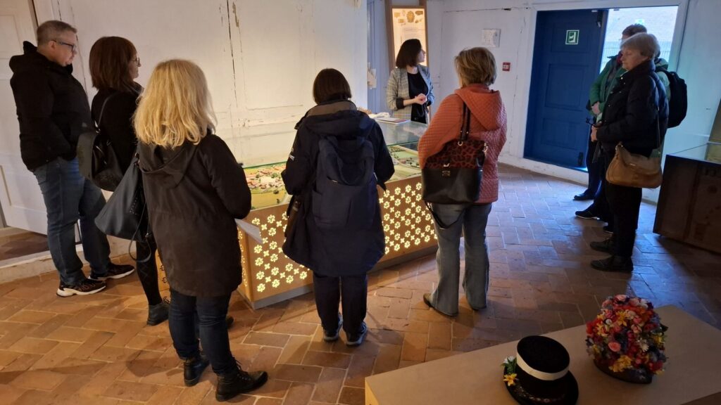 Uczestnicy oprowadzania oglądają makietę w gablocie ustawionej na środku sali wystawowej, podczas gdy przewodniczka omawia szczegóły prezentacji (kliknięcie powiększy zdjęcie).