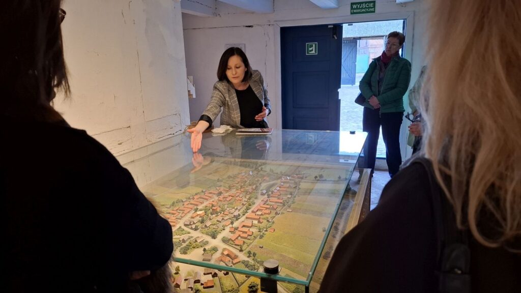 Przewodniczka wskazuje elementy makiety miasta w gablocie, a stojący wokół zwiedzający obserwują i słuchają jej objaśnień (kliknięcie powiększy zdjęcie).