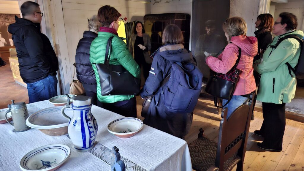 Grupa zwiedzających stoi w historycznej izbie przy stole nakrytym ceramicznymi naczyniami, słuchając przewodniczki opowiadającej o ekspozycji (kliknięcie powiększy zdjęcie).
