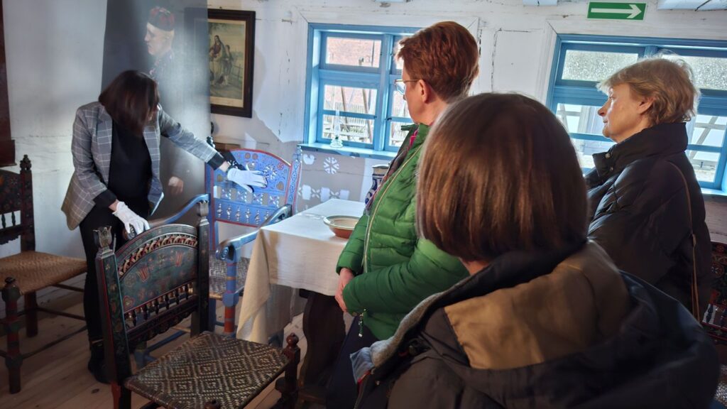 Przewodniczka pokazuje krzesło zdobione w jamneńskie wzory stojące przy stole w muzealnym wnętrzu, a uczestnicy oprowadzania obserwują prezentowany eksponat (kliknięcie powiększy zdjęcie).