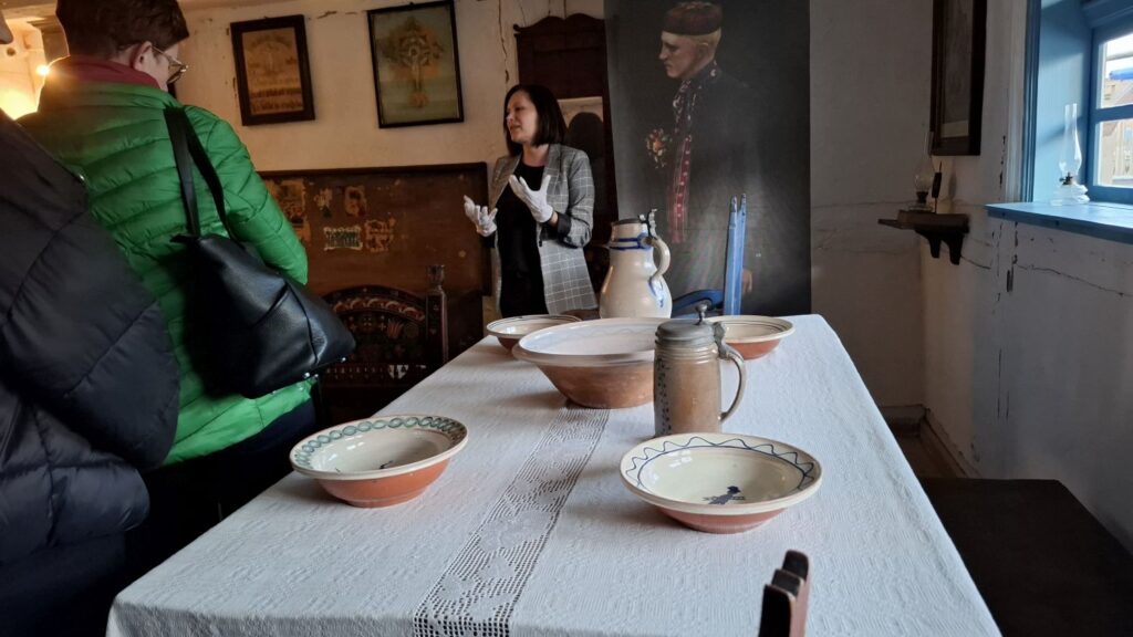 Zwiedzający stoją przy długim stole nakrytym naczyniami, słuchając przewodniczki opowiadającej o wyposażeniu dawnej izby (kliknięcie powiększy zdjęcie).