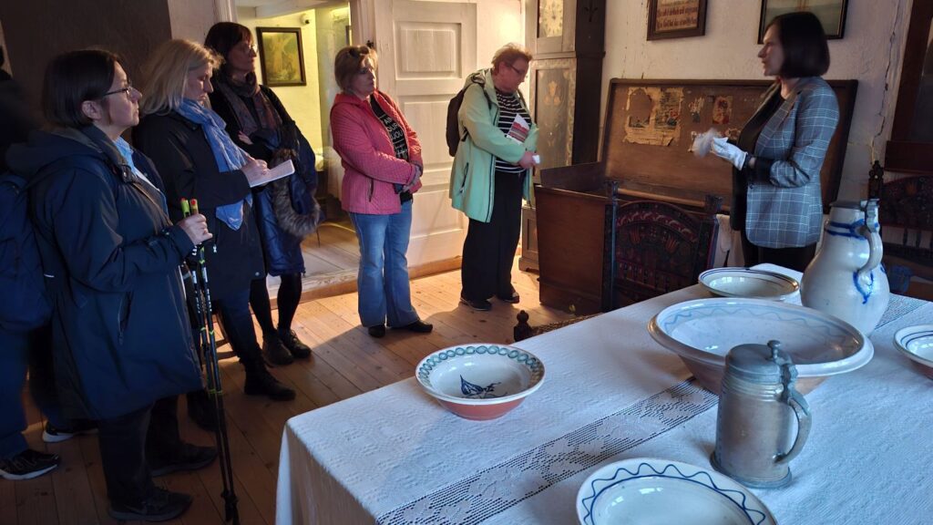 Przewodniczka opowiada o dawnym wyposażeniu domu przy otwartej skrzyni, a zwiedzający stoją wokół i jej słuchają (kliknięcie powiększy zdjęcie).