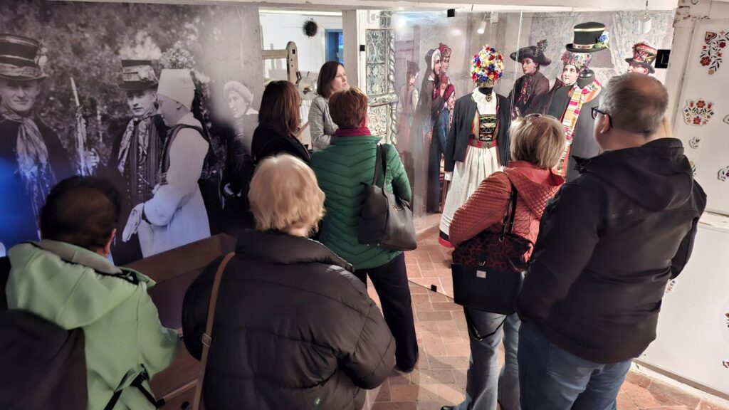 Grupa zwiedzających ogląda ekspozycję z manekinami w tradycyjnych strojach ludowych oraz archiwalne fotografie na ścianie (kliknięcie powiększy zdjęcie).