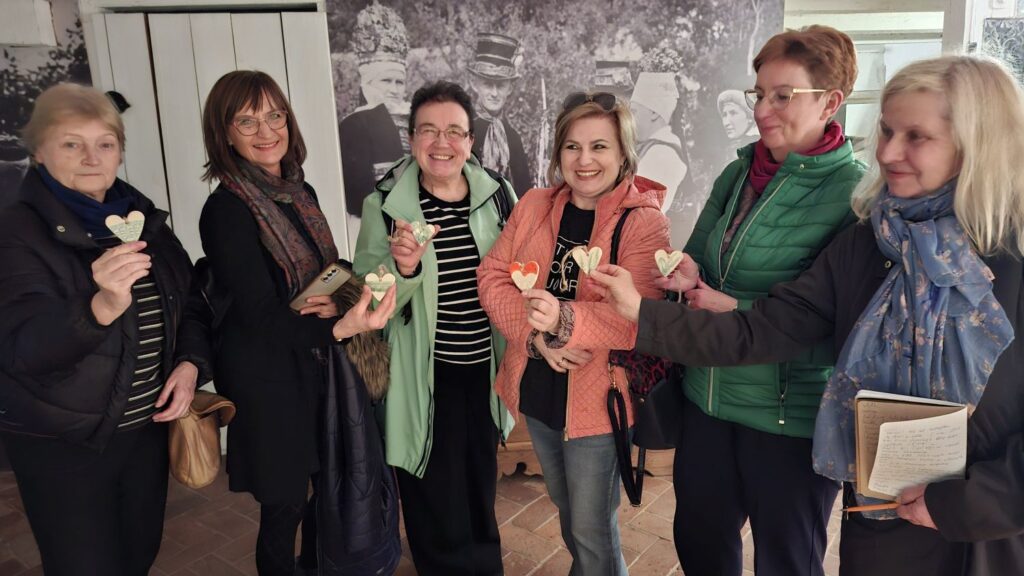 Kilka kobiet stojących w sali wystawowej trzyma w dłoniach małe serduszka i uśmiecha się do zdjęcia na tle ekspozycji (kliknięcie powiększy zdjęcie).
