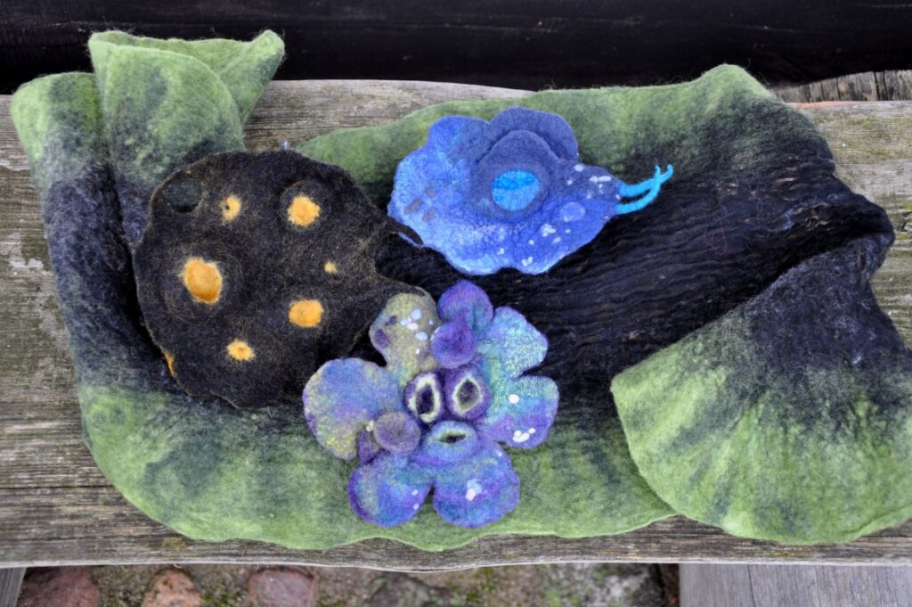 Zestaw filcowanych prac z wełny ułożonych na drewnianej ławce – kwiaty, etui oraz dekoracyjne elementy w różnych kolorach (kliknięcie powiększy zdjęcie).