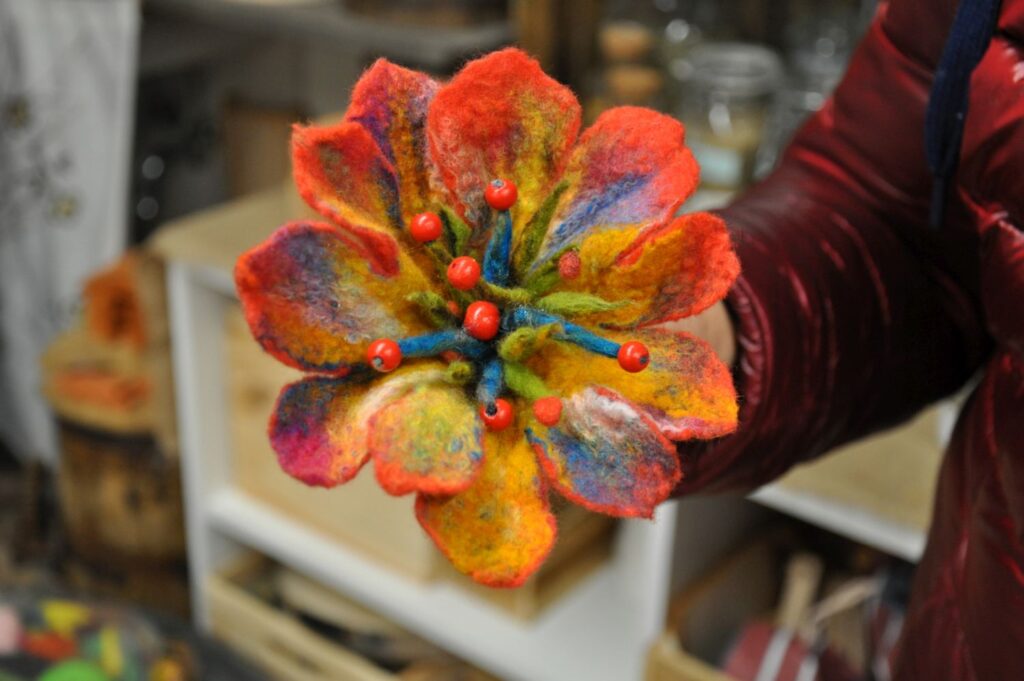 Kolorowy kwiat wykonany z filcowanej wełny, ozdobiony koralikami, trzymany w dłoni uczestniczki warsztatów (kliknięcie powiększy zdjęcie).